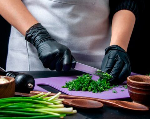 Una dona amb guants negres talla verdures en una taula de tallar.